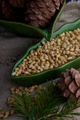 Lot of peeled pine nuts in a green plate in the shape of a leaf on burlap. Cedar cones and a...