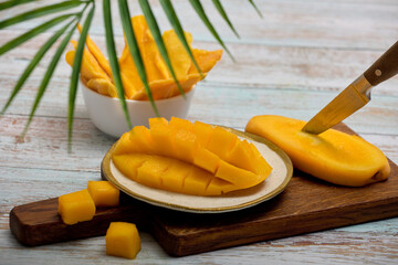 Sweet tropical mango is cut into cubes and laid out on a porcelain saucer with a gilded border. The delicious, juicy, healthy flesh of a ripe mango is cut with a sharp knife on a wooden board.