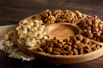 Dried cashews, almonds, hazelnuts and walnuts are laid out on a wooden rack. Wooden round plate with various types of nuts, standing on a burlap on the table. 