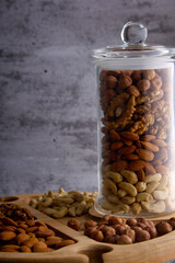 On a wooden plate there is a transparent glass jar with a lid filled with assorted nuts. Vertical photo of walnuts, cashews, almonds and hazelnuts in a jar