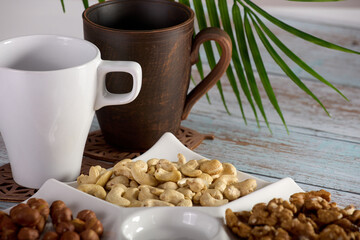 Mix of hazelnuts, almonds, cashews and walnuts is laid out in a white bowl standing on a wooden table. Assorted dried nuts of various types are laid out in close-up on a white square plate. 