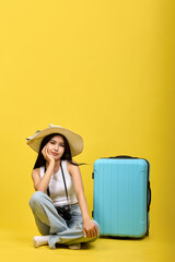 A wise Asian girl in a summer hat holds her head with her hand, sits next to a suitcase. Asian...