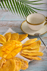 Vertical shot of sliced mango on a white platter along with dry fruit plates. On a wooden table, an au pair on a background of palm leaves and tropical mango on an irregular porcelain plate