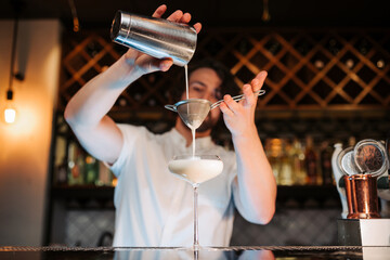 A skilled male bartender expertly pours a fresh cocktail into a martini glass, showcasing his...