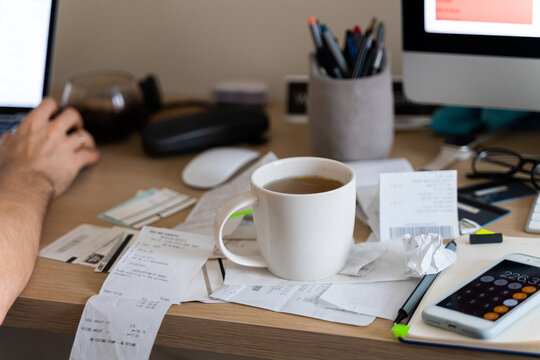 Man works on messy desk with cup on top of receipt, invoice, tax forms