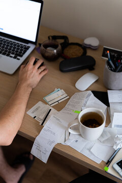 Man works on messy desk with cup on top of receipt, invoice, tax forms