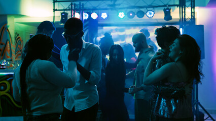 Romantic people dancing in pairs on dance floor, partying with slow music at discotheque. Cheerful couples enjoying dance party with stage lights and drinks, entertainment event. Handheld shot.