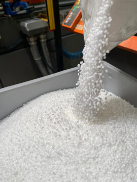 pouring of a bag of white plastic pellets on a gray plastic drawer, production of plastic parts, plastic injection, spilling of plastic pelets, with a plastic injector out of focus in the background, 