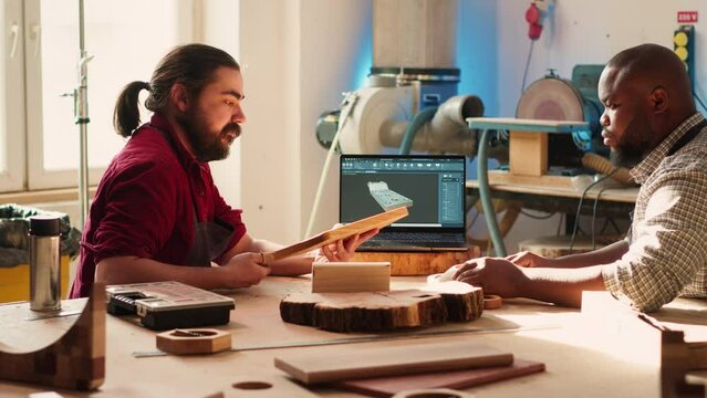 Artisan and apprentice comparing wood art piece with schematic displayed on CAD software on laptop. Woodworking experts in studio crosschecking wooden object with notebook blueprint, camera A