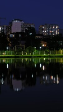 Timelapse, vertical slider video: apartment residential buildings facades with blinking illuminated windows with reflection in the water surface in Batumi at night. People are walking fast: time lapse