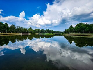 Tranquil lake surrounded by lush greenery and blue sky with fluffy white clouds reflecting on the...