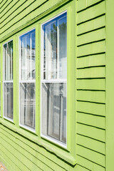 A lime green colored wooden clapboard exterior wall of a house with three single hung vintage glass windows. White lace curtains are hanging in the windows. The narrow trim on the window is white.