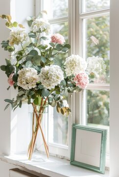 Pink and white hydrangeas in a  vase on table, a small green picture mockup frame, against the window 
