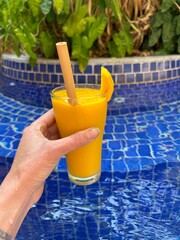 hand holding bright yellow tropical mango drink in front of pool - travel texture in Cambodia