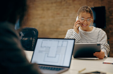 Smiling female freelancer in eyeglasses is talking phone with client while sitting in coworking