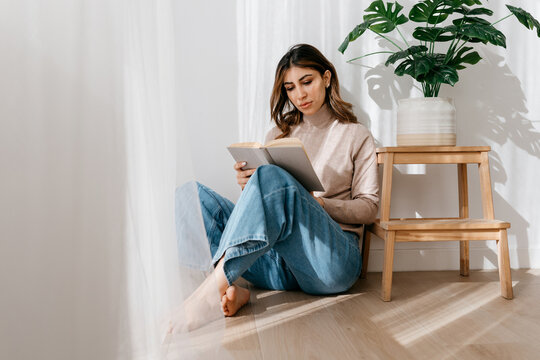 Woman reading book while sitting near stool with plant