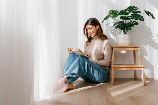 Cheerful woman reading book on floor in apartment