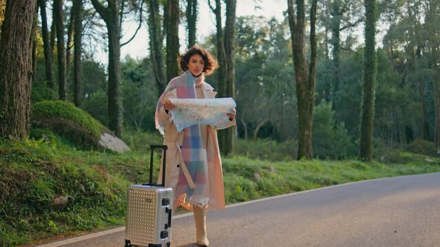 Girl tourist studying map on scenic forest roadside with suitcase. Woman journey