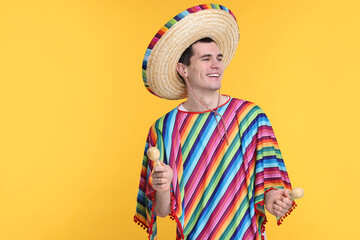 Young man in Mexican sombrero hat and poncho with maracas on yellow background. Space for text