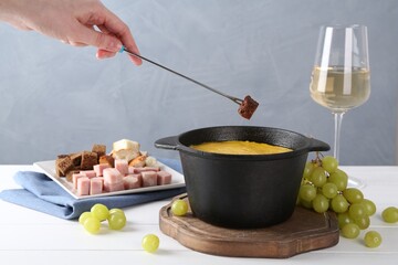 Woman dipping piece of bread into fondue pot with tasty melted cheese at white wooden table against...