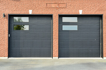 Exterior of a red brick residential building with double black metal car garage doors. There are two small glass panels in each solid door. The driveway is concrete. There's a lit fixture on the wall.