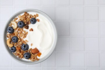 Bowl with yogurt, blueberries and granola on white tiled table, top view. Space for text