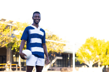African American young male athlete holding a rugby ball, standing on field outdoors, copy space