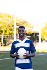 African American young male athlete holding a rugby ball, standing on field outdoors, copy space