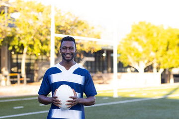 African American young male athlete holding rugby ball, standing on field outdoors, copy space
