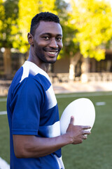 African American young male athlete holding a rugby ball, smiling at the camera on the field outdoor