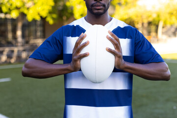African American young male athlete holding a rugby ball, standing on field outdoors