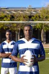 Two African American young male athletes, holding a rugby ball, standing on field outdoors