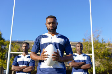Three African American young male athletes holding a rugby ball, standing on field outdoors
