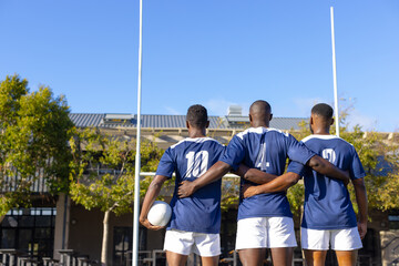 Three African American young male athletes wearing blue jerseys, standing on a rugby field outdoors