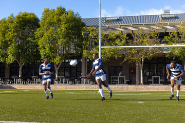 Three African American young male athletes in blue and white gear play rugby on field outdoors