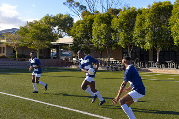 Three African American young male athletes wearing sportswear are playing rugby on field outdoors