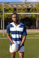 African American young male athlete holding rugby ball on field outdoors, looking serious