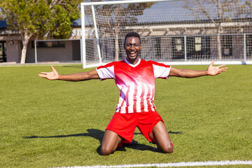 African American young male athlete kneeling on soccer field outdoors, arms wide, celebrating
