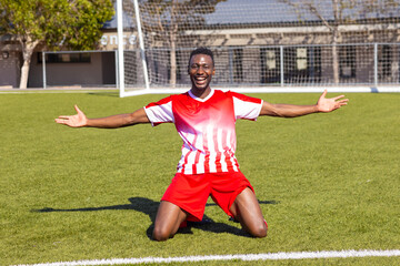 African American young male athlete kneeling on soccer field outdoors, arms wide open