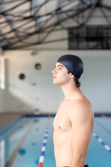 Caucasian young male swimmer standing by the pool indoors, looking thoughtful