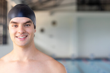 Caucasian young male swimmer standing by pool indoors, wearing cap, smiling, copy space