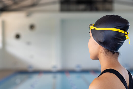 Biracial young female swimmer wearing a black cap is looking at a pool indoors, copy space