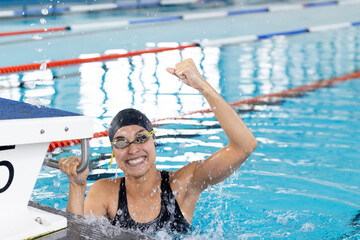 Biracial young female swimmer celebrating indoors in pool, wearing goggles