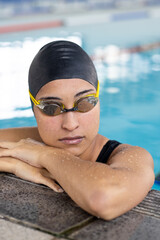 Biracial young female swimmer resting at pool edge indoors, looking thoughtful