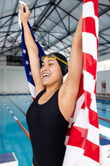 Young biracial female swimmer celebrating with American flag by poolside indoors