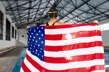 Young biracial female swimmer holding American flag by poolside indoors