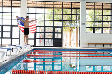 Caucasian young male swimmer holding American flag by poolside indoors, copy space