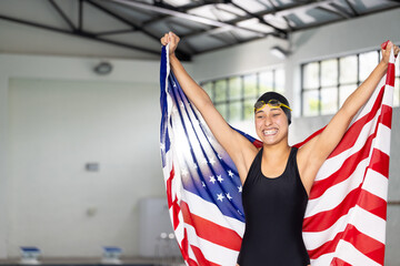 A biracial young female swimmer is holding an American flag indoors, beaming with pride, copy space