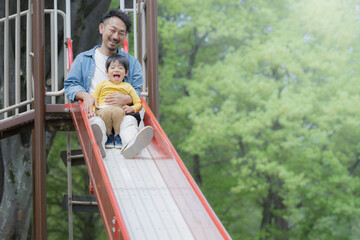 Obraz premium 新緑の公園の滑り台で遊ぶアジア人（日本人）の親子 2人家族 パパ(父親）の子育てや育児の休日のイメージ