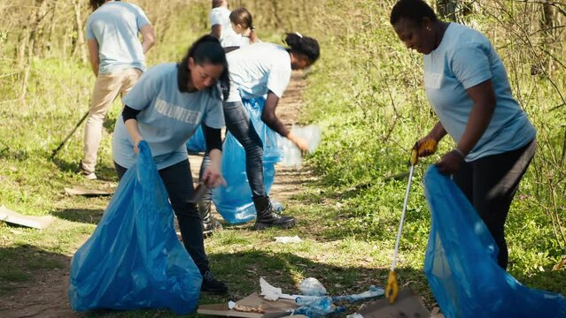 Group of diverse volunteers collecting rubbish from the woods and recycling in a garbage disposal bag, litter cleanup responsibility. Ecology activists picking up trash and plastic waste. Camera A.
